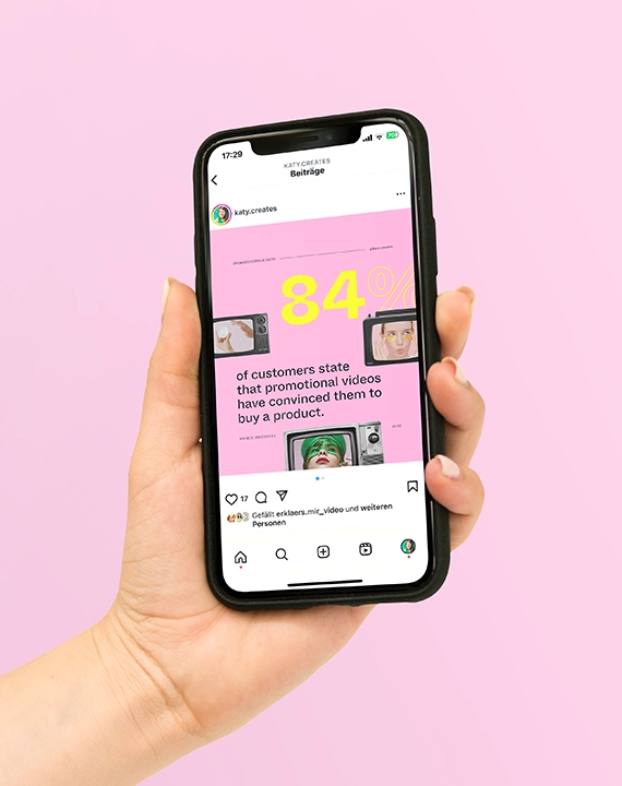 Eine Hand hält ein schwarzes Iphone vor einem rosa Hintergrund in die Höhe