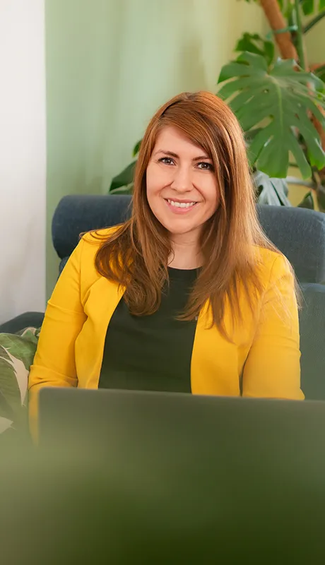 Kathrin Schönborn schaut in die Kamera, sie trägt einen gelben Blazer, sitzt auf einem blauen Sofa und arbeitet am Laptop