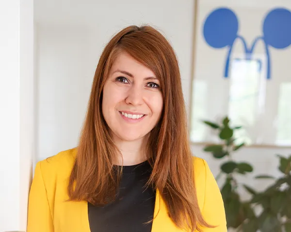 Kathrin Schönborn trägt einen gelben Blazer, steht in ihrem Büro und lächelt in die Kamera